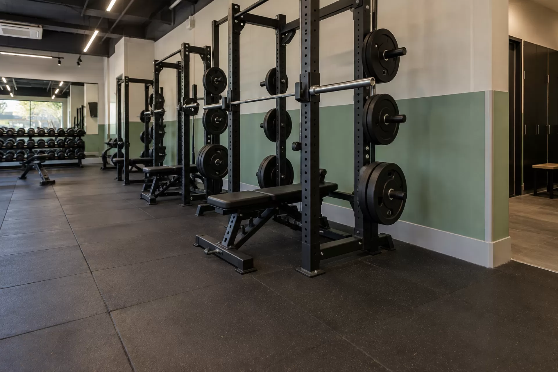 Sala de musculação com racks e bancos e proteção instalada na parede atrás dos equipamentos